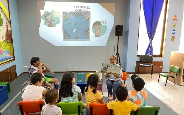 Grupo de niños y niñas frente a una pantalla proyectada en la pared y a una mujer que enseña un libro