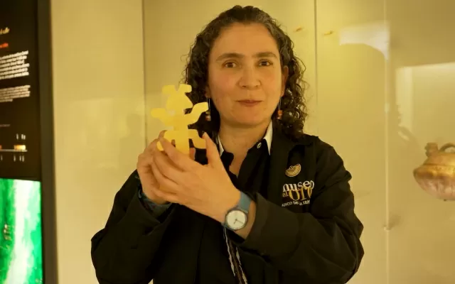 Una mujer de cabello negro, usando ua chaqueta negra del museo del oro, sosteniendo una obra con sus manos