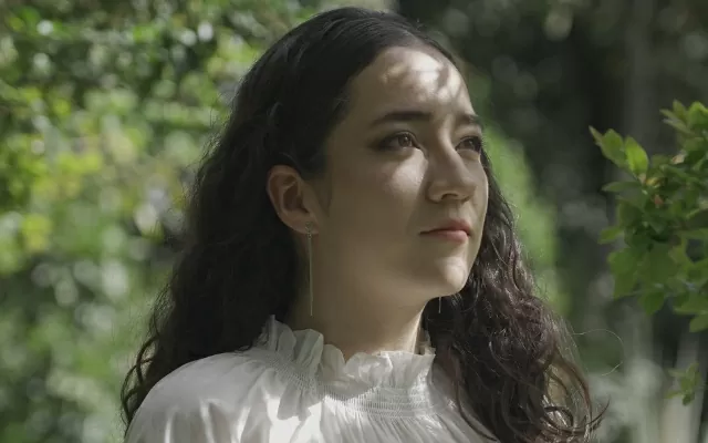 Mujer joven, de cabello negro, con blusa blanca, rodeada de naturaleza, mirando al horizonte