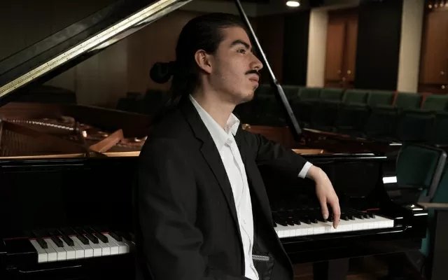 Hombre de cabello negro y largo, recogido, usando traje negro, sentado junto a un piano, posando con un brazo sobre él.