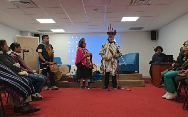 Un hombre y una mujer, ubicados en el centro de un auditorio, rodeados de otras personas, y con una proyección de pantalla a sus espaldas.  