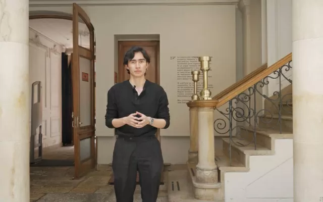 Hombre, vestido de negro, hablando y moviendo sus manos, ubicado junto a unas escaleras, 