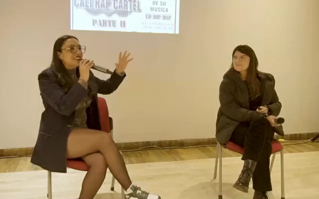 Dos mujeres, sentadas, sosteniendo un microfono, con una imagen proyectada a sus espaldas