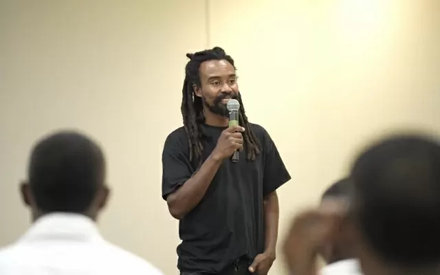 Hombre, de cabello largo y oscuro, usando camiseta negra, sonriendo levemente, sosteniendo un microfono con su mano, y hablando hacia el público 