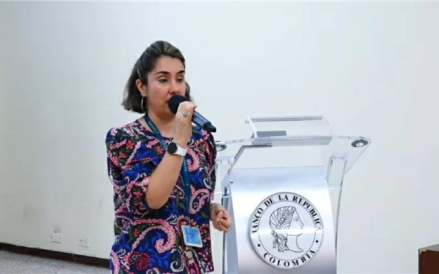 Una mujer, de cabello corto, usando una blusa de colores, sosteniendo un microfono con su mano, mientras se dirige al público 