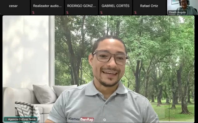 Hombre, usando gafas, y una camiseta gris tipo polo de Banrep, con un fondo de naturaleza, mientras participa en una videoconferencia