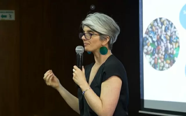 Mujer, usando una blusa negra, sosteniendo un micrófono, dando una conferencia 