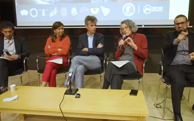 Dos mujeres, y tres hombres, sentados alrededor de una mesa, con una pantalla al fondo, dos de ellos sostienen un micrófono, mientras imparten una conferencia 