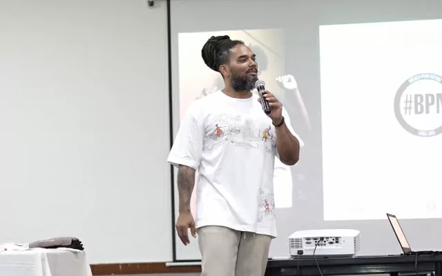 Hombre, de cabello largo y recogido, usando camiseta blanca, sosteniendo un microfono, mientras se dirige al público 