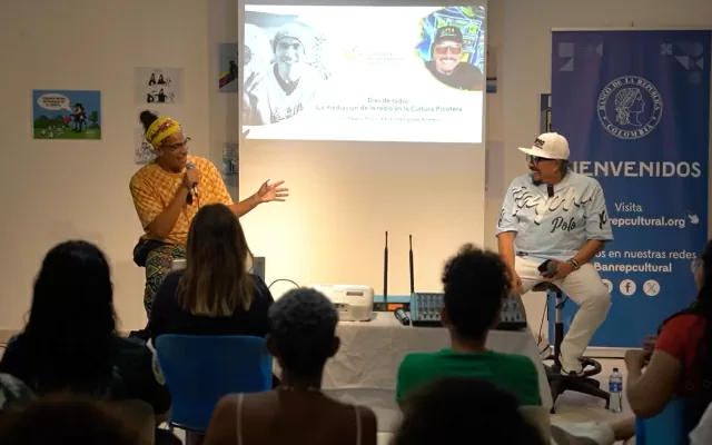 Dos hombres, sentados, sosteniendo un micrófono, dando un conversatorio frente a un público 