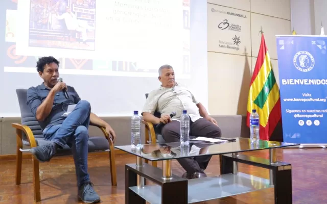 Dos hombres sentados, alrededor de una mesa de vidrio, sosteniendo un micrófono con sus manos, con una pantalla a sus espaldas