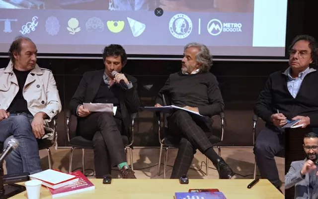 Cuatro hombres, sentados alrededor de una mesa, sosteniendo un micrófono, dando un conversatorio