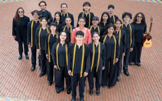 Diecinueve músicos en plano completo vestidos de negro y una música vestida de naranja, de pie, mirando fijamente