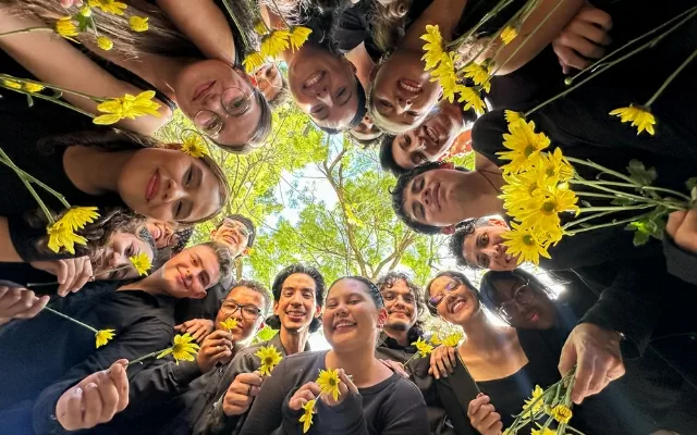 Grupo de mujeres y hombres, vestidos de negro, sonriendo, con una flor amarilla en la mano 