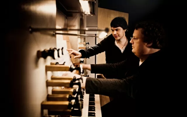 Dos músicos, vestidos de negro, mirando una partitura, frente a un órgano 