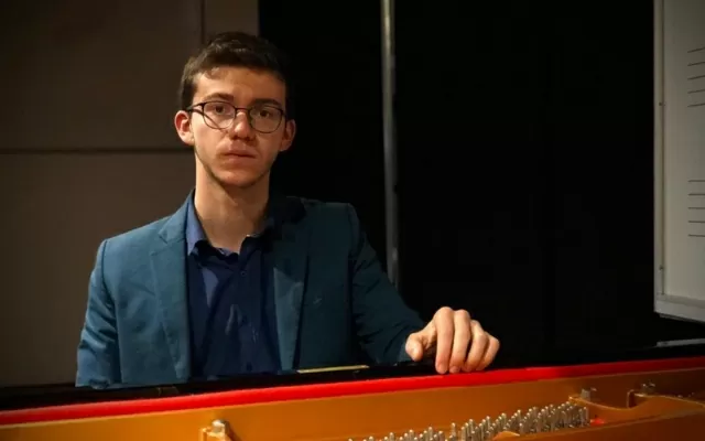 Hombre usando gafas, con un traje azul oscuro, y una mano sobre el piano