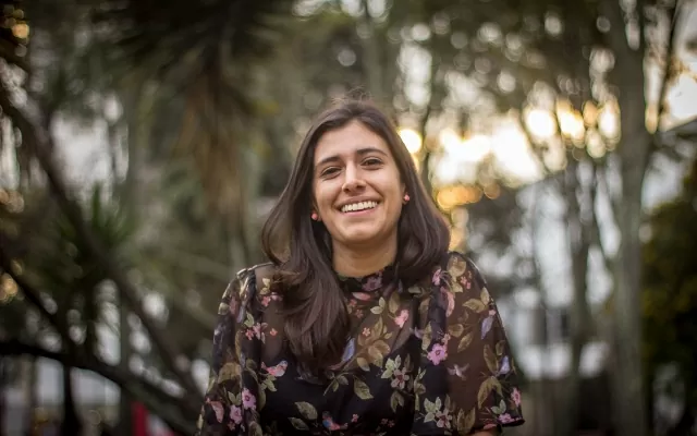 Mujer de cabello negro, sonriendo, usando una blusa de flores, con un fondo de naturaleza 