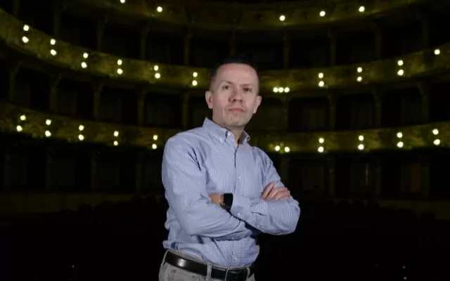 Hombre usando camisa azul, con los brazos cruzados, con un auditorio como fondo