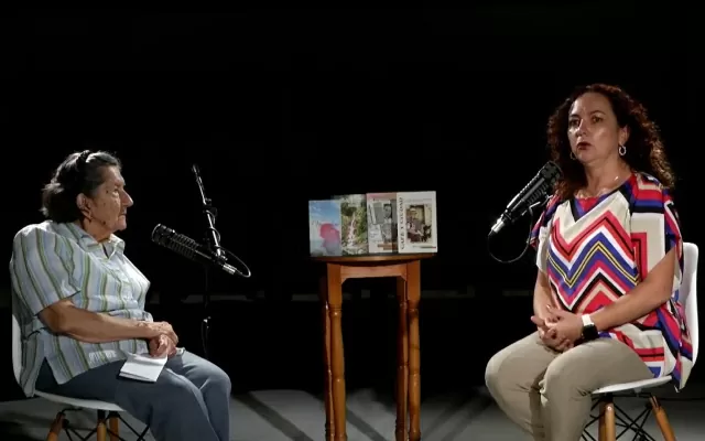 Dos mujeres, sentadas, alrededor de una mesa con libros, cada una con un micrófono en frente