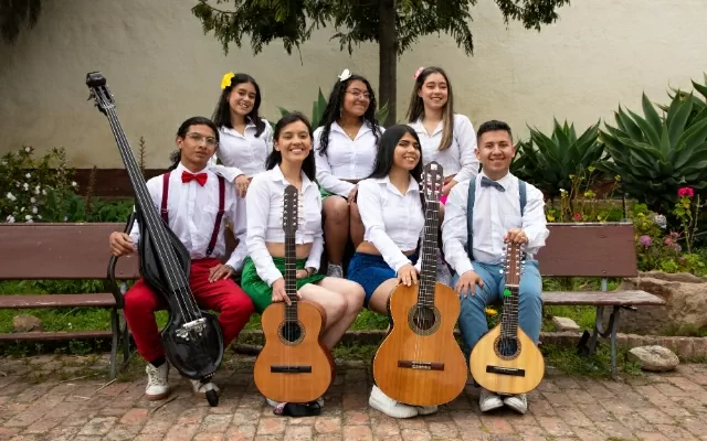 Cinco mujeres y dos hombres, sentados sobre un banco café al aire libre, sosteniendo cuatros instrumentos