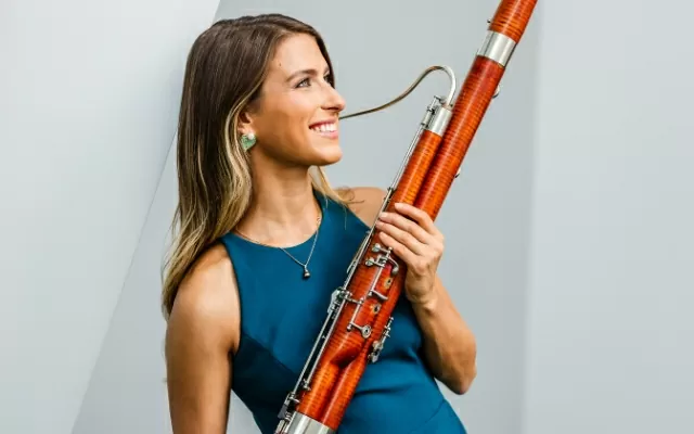 Música sonriendo, usando una blusa azul, mientras mira en otra dirección y sosteniendo un fagot