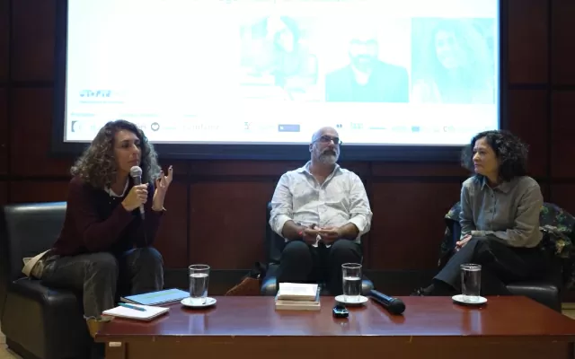 Un hombre y dos mujeres, sentados, mientras una de ellas sostiene un micrófono, y se dirige al público 