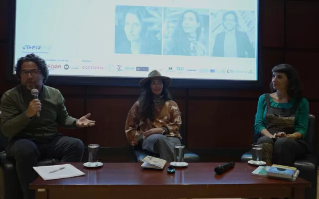 Dos mujeres, un hombre, sentados al rededor de una mesa, mientras uno de ellos sostiene un micrófono y habla a los asistentes