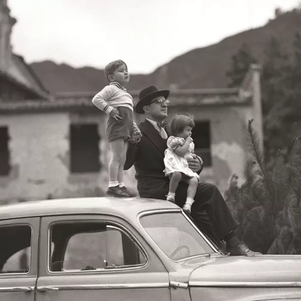 Fotografía a blanco y negro de un padre junto a sus hijos, el junto a su hijo más pequeño reposan sobre el techo de un automóvil y el más grande se para sobre el techo del mismo