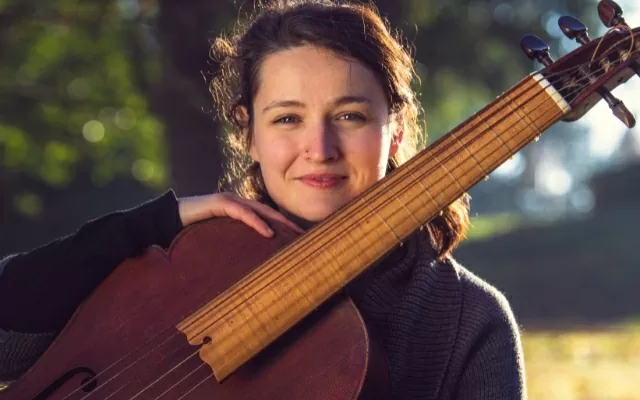 Mujer sonriendo, al aire libre, rodeada de naturaleza, en plano medio corto, sosteniendo una viola da gamba