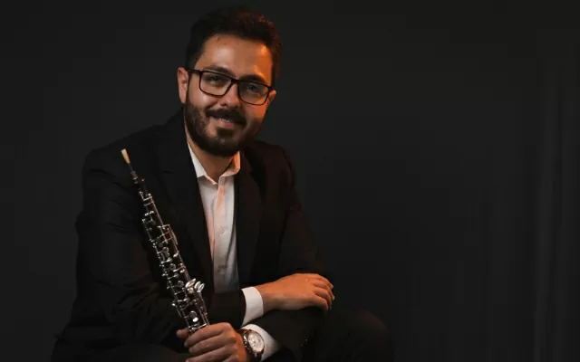Retrato de músico sentado, usando gafas y traje negro, con fondo negro, sosteniendo un oboe con su mano