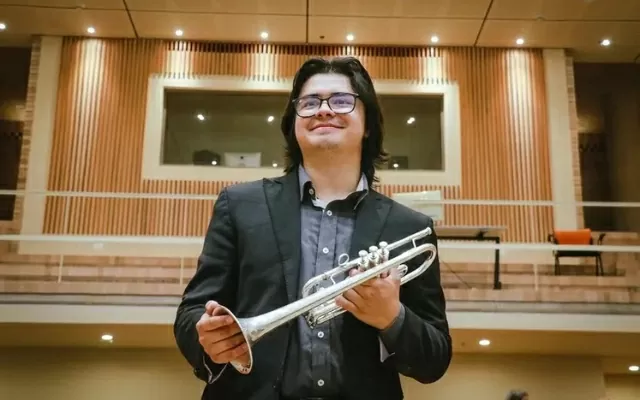 Hombre usando un traje negro y gafas, sonriendo, con una trompeta en la mano, mientras mira en otra dirección