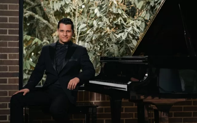 Músico usando un traje negro, sonriendo y mirando fijamente, sentado junto a un piano negro
