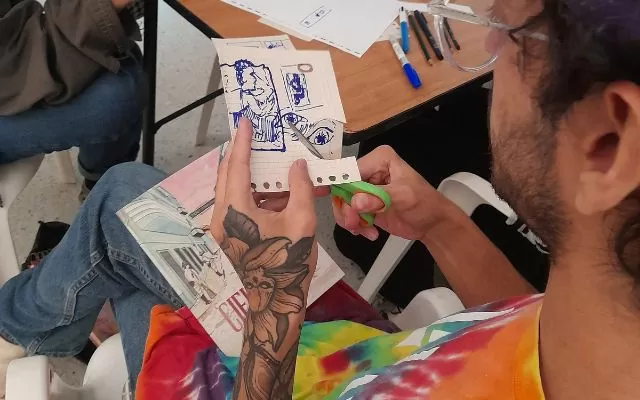 Foto de un hombre de camisa de colores, gafas, que está sentado con unas tijeras en la mano, recortando un dibujo; el hombre lleva un tatuaje grande en su brazo izquierdo 