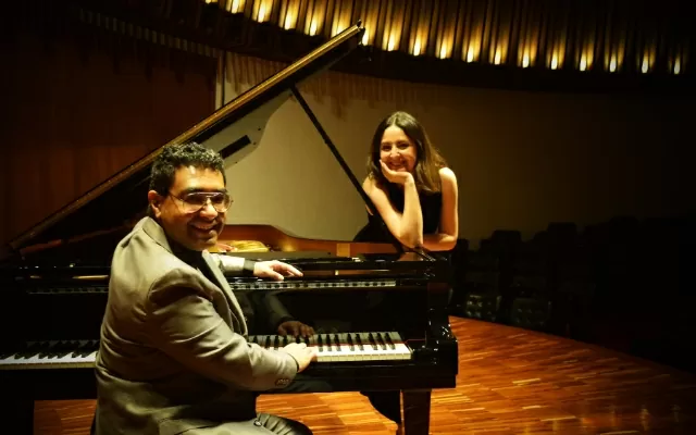 Dos músicos alrededor de un piano negro, en un auditorio, mirando fijamente y sonriendo