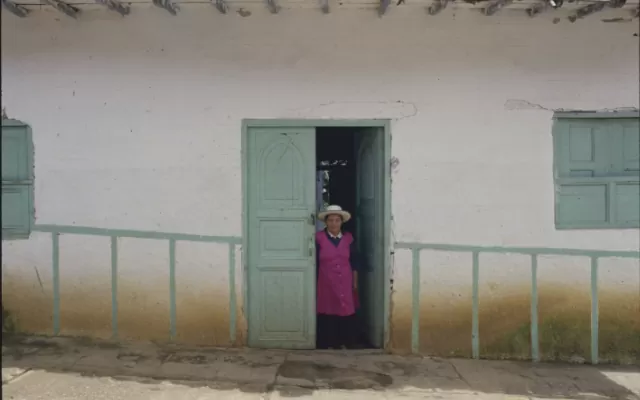 Vivienda con paredes blancas de Pachavita. Las puertas y ventanas azules de madera hacen juego con la falsa baranda pintada sobre la pared. En el centro, una mujer campesina con sombrero posa para la fotografía.