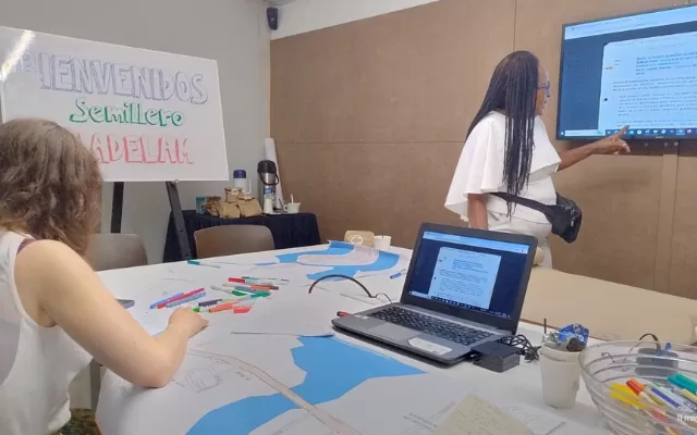 Taller participativo en una sala. Personas alrededor de una mesa con materiales mientras una mujer señala una pantalla