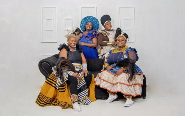 Imagen a color de dos mujeres sentadas en un sofá negro, dos mujeres de pie, sonriendo, mirando fijamente  y con un fondo blanco