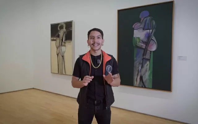 Hombre, usando chaleco negro, moviendo sus manos mientras habla, con dos obras de Luis Caballero al fondo 