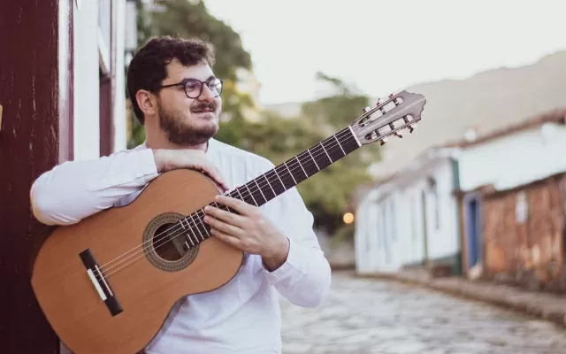 Músico usando una camisa blanca, al aire libre, en plano medio, observando al horizonte mientras sostiene una guitarra 