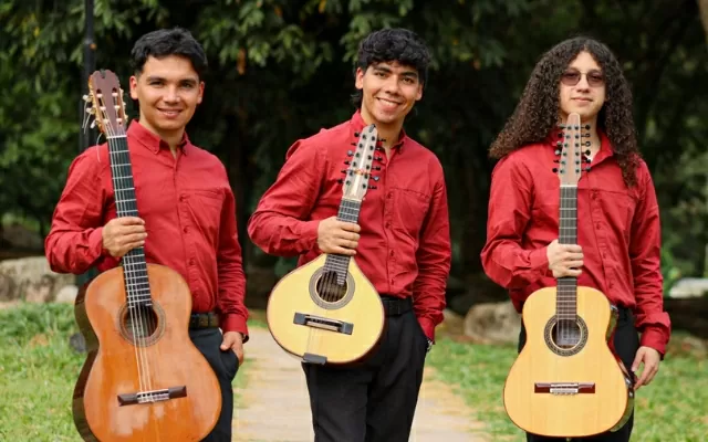 Tres artistas, sonriendo, usando camisa roja, sosteniendo sus instrumentos y posando al aire libre