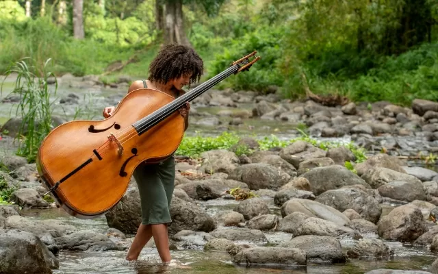 Artista caminando a través de un río, mientras observa sus pies y sostiene un instrumento