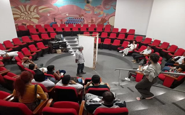 Imagen de sala de música, que tiene muebles rojos, está organizado en forma circular y hay varias personas escuchando una conversación 