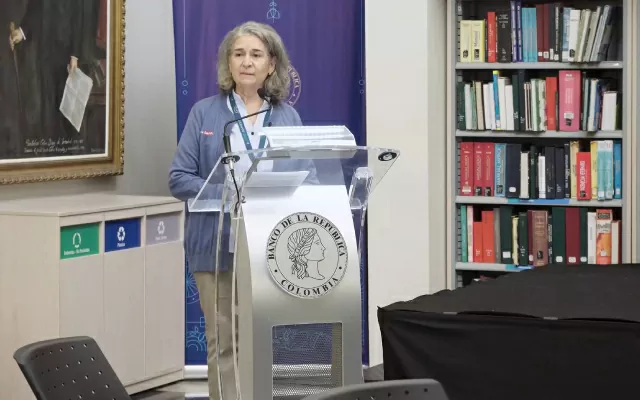 Mujer, usando saco azul y pantalón beige, hablando frente a un micrófono, junto a una biblioteca 