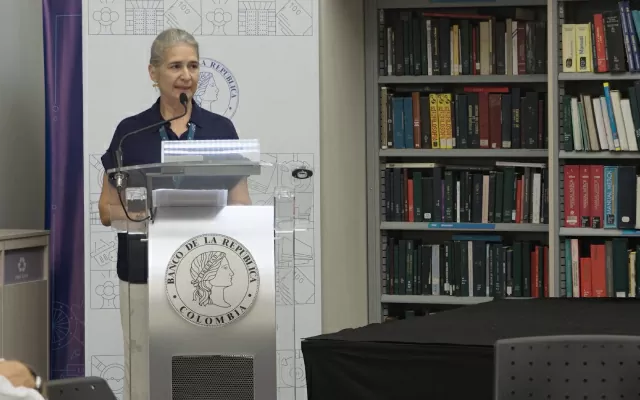 Mujer, con el cabello recogido, usando una blusa azul oscura, hablando frente a un micrófono, con una librería a su lado 