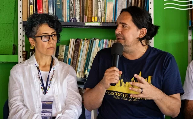Hombre hablando con micrófono y mujer a su lado escuchando con biblioteca de fondo