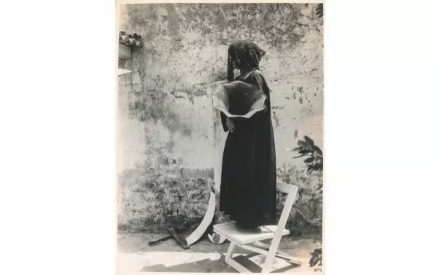 Fotografía en blanco y negro donde se ve una mujer vestida de negro, parada sobre una silla blanca