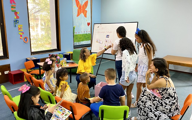 Grupo de niños y niñas sentados alrededor de la tallerista y de un tablero, donde los participantes señalan una imágenes