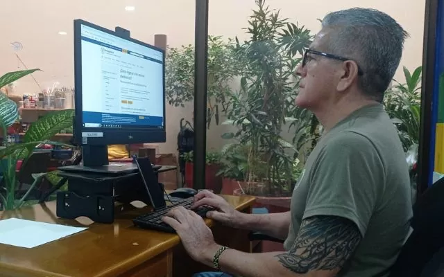Se ve un señor mayor, que mira fijamente su computador mientras en la pantalla aparece información del portal de Banrepcultural sobre recursos electronicos. El señor se encuentra en una oficina, rodeado de elementos propios del lugar y algunas plantas verdes.