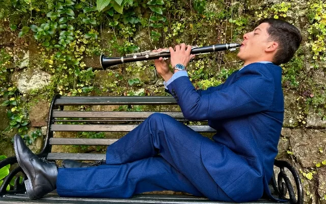 Retrato a color de Víctor Suárez, usando un traje azul, sentado en un banco, tocando el clarinete 