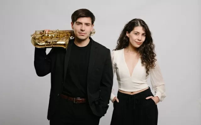 Hombre usando traje negro, sosteniendo un saxofón sobre su hombro, mujer con las manos en sus bolsillos, ambos en plano medio